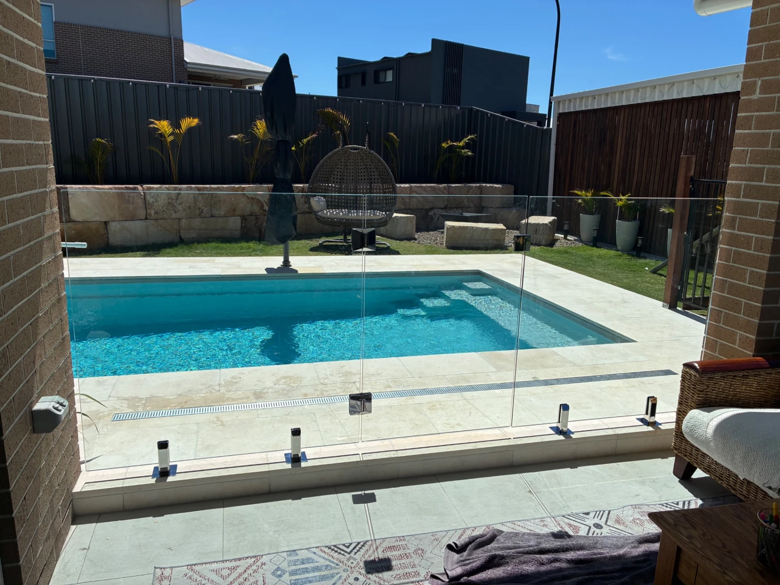 Frameless glass fence on stainless-steel spigots around a blue pool beside a paved patio