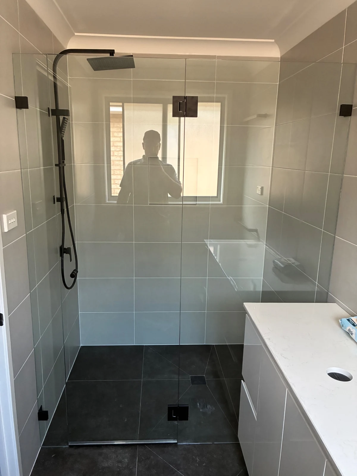 Frameless glass shower screen with black hinges and handle in a grey-tiled bathroom