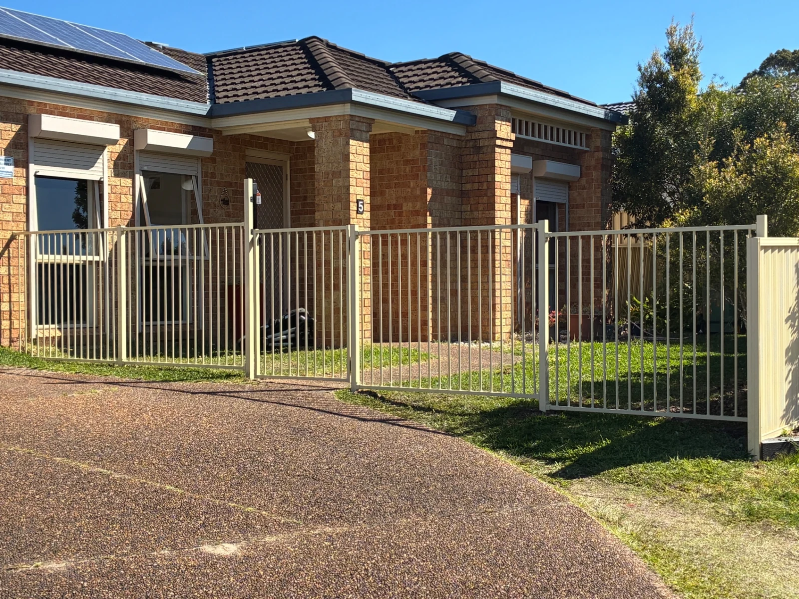 Light-coloured metal picket boundary fence and gate in front of a brick home