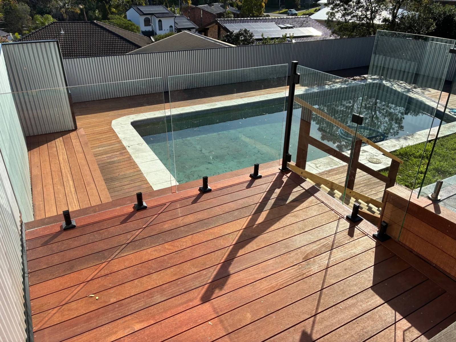 Frameless glass pool fence with black gate hardware beside a reddish timber deck
