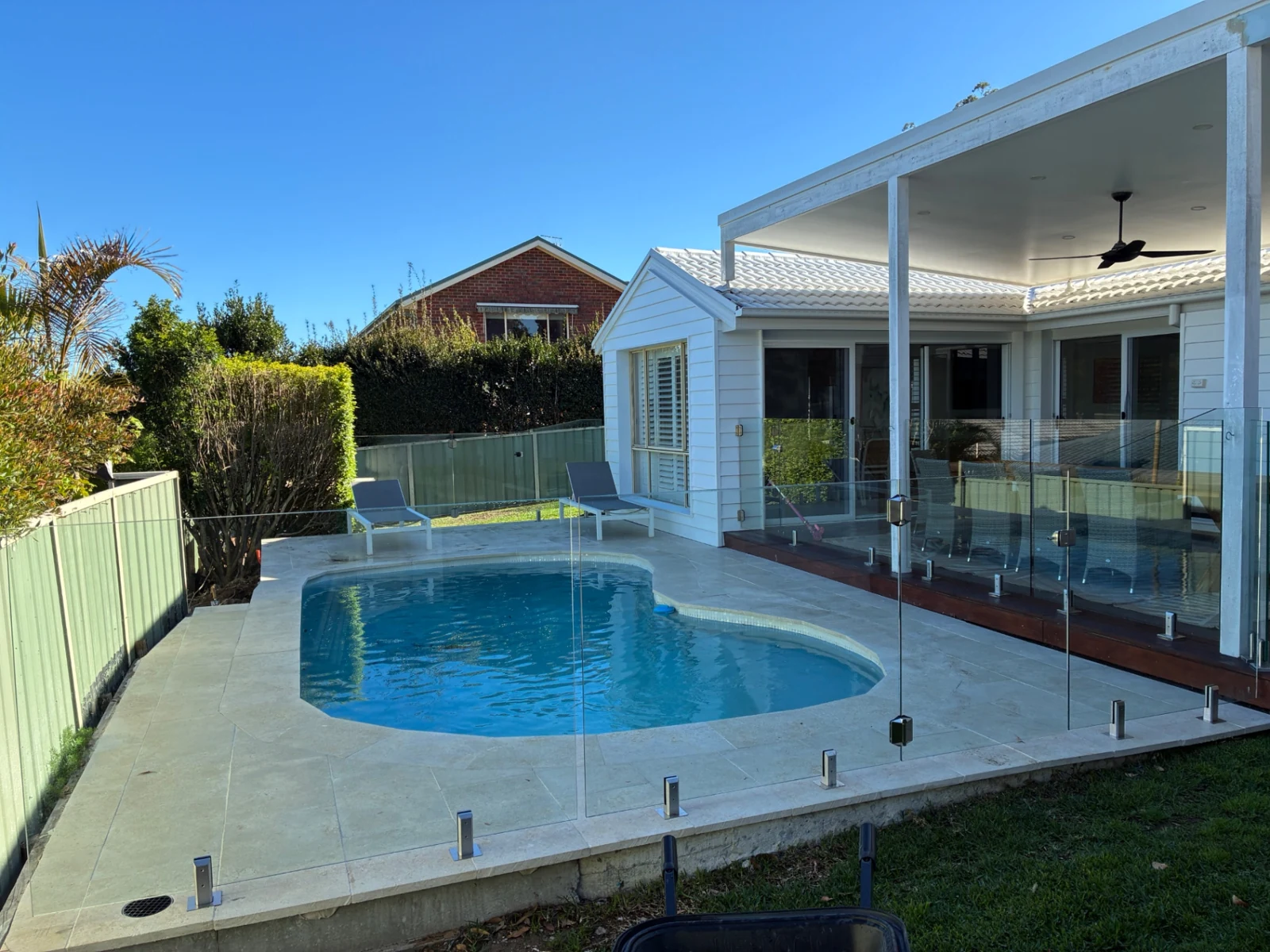 Clear glass fence around a curved blue swimming pool beside a white two-storey home